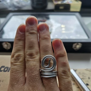 Handgjord Silvrig spiralring i metall - Handgjord ring i silvrig metall med cool spiraldesign som ger en edgy och modern vibe. Ringen består av flera varv och har en öppen, konstnärlig form som sticker ut på handen. Perfekt för dig som gillar statement-smycken och vill ha något annorlunda. Ringen är I aluminium och i st 18.5 mm