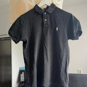Svart slim fit piké från Polo Ralph Lauren - Svart pikétröja från Polo Ralph Lauren i slim fit-modell. Klassisk krage med två vita knappar och broderad logga på bröstet. Tillverkad i mjuk bomull, perfekt för en clean och stilren look. Kortärmad och passar dig som gillar enkel men snygg design.
