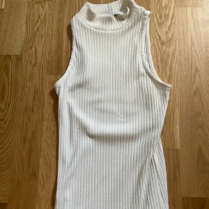 Vit ribbad ärmlös polotopp - Säljer en stilren vit polotopp i ribbat material. Toppen är ärmlös med hög hals och har en tight passform som ger en clean och modern look. Perfekt att styla med jeans eller kjol för en fräsch vibe.