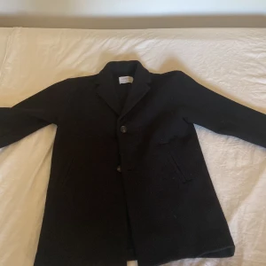 Svart kappa från Zara, stl 140 - Stilren svart kappa från Zara med klassisk krage och knäppning framtill. Jackan har två fickor och är helsvart, perfekt för dig som gillar en clean look. Passar dig som vill ha en enkel men snygg jacka till höst och vinter.