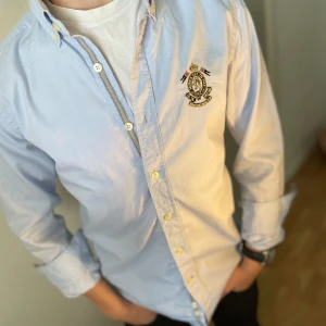 Ljusblå skjorta från Morris - Snygg ljusblå skjorta från Morris med broderad logga på bröstet. Klassisk krage, knäppning framtill och långa ärmar. Skjortan är i bomull och har en stilren, tidlös look som passar perfekt till jeans eller chinos.                                              Osäker på priset så kom med förslag!!!