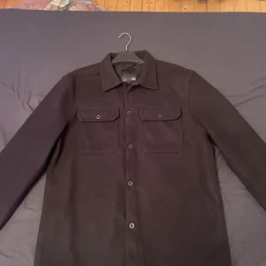 Svart overshirt från 157, storlek L - Snygg svart overshirt från 157 i storlek L passar även dom som har storlek M. Jackan har klassisk skjortkrage, två bröstfickor med knappar och hel knäppning framtill. Materialet är mjukt och känns som ull, perfekt för lager-på-lager. Enkel och stilren design som funkar till många outfits.