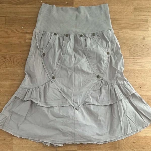 Grå långkjol från Mamalicious - Säljer en grå långkjol från Mamalicious med bred ribbad midja och snygga knappdetaljer framtill. Kjolen har volangkant och är tillverkad i mjuk bomull med polyester. Perfekt för dig som gillar bekväma och stilrena plagg.