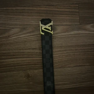  svart rutigt bälte - Säljer ett stilrent  bälte i svart med klassiskt rutigt mönster och en snygg guldfärgad -spänne. Bältet är tillverkat i skinn och har en modern, rak form som passar perfekt till jeans eller kostymbyxor.