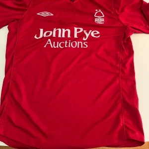Röd Nottingham Forest fotbollströja Umbro - Säljer en röd Nottingham Forest fotbollströja från Umbro i storlek S. Tröjan har korta ärmar, V-ringad krage med meshdetaljer och tryck i vitt med klubbmärke och sponsor. Materialet är lätt och andas, perfekt för match eller träning.