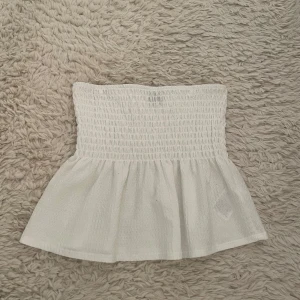 Vit tubtopp från Gina Tricot - Supersöt vit bandeau tubtopp från Gina Tricot med smockad överdel och volang nertill. Perfekt för varma dagar och enkel att styla med jeans eller kjol. Toppens struktur ger en trendig och luftig känsla.