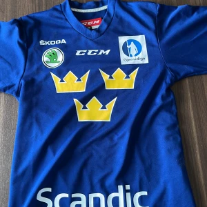 Blå Tre Kronor fotbollströja CCM - Säljer en blå fotbollströja från CCM med Tre Kronor-tryck i gult på bröstet och flera sponsorer som Scandic, Skoda och Gjensidige. Tröjan har korta ärmar, V-ringad krage och är gjord i ett lätt, ventilerande syntetmaterial. Perfekt för dig som gillar landslagskänsla.