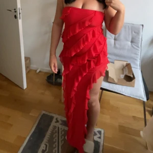 Röd klänning med volang - Säljer en röd axelbandslös långklänning med volangdetaljer i flera lager och slits på ena sidan . Den är beställd ifrån oh polly och endast använd en gång under min bal. Inga defekter eller skador. Perfekt för bal eller annan tillställning. 
