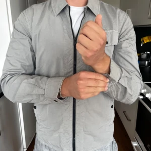 Grå vindjacka med dragkedja - Snygg grå vindjacka med klassisk krage och svart dragkedja framtill. Jackan har en bröstficka med knapp och två sidofickor. Lätt och smidig modell som passar perfekt till vår och höst. Långärmad och i ett tunt, skönt polyestermaterial. Nypris 3200