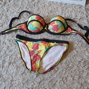 2000-tals bikini - Färgglad bikini med blommigt mönster i rött, grönt och orange på gul botten. Överdelen har vadderade kupor och svarta kanter, underdelen har matchande blommigt tyg och svarta detaljer i midjan. Perfekt för strandhäng och poolparty.