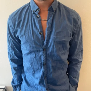 Blå skjorta från Filippa K, M - Snygg blå skjorta från Filippa K i storlek medium. Skjortan har klassisk krage med knappar, bröstficka och långa ärmar. Tillverkad i mjuk bomull och har en normal passform. Perfekt för dig som gillar stilrena och enkla plagg.