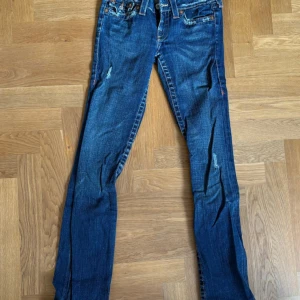 True Religion blå jeans bootcut strl.26 - Mörkblå True Religion jeans med low waist och lätt bootcut, Billy, detaljerade fickor bak och slitningar fram. OBS jeansen är i fint skick men dragkedjan är trasig, kostar ej mycket att byta! Tillverkade i USA, strl.26. 36,5cm rakt över och innerben:86 cm