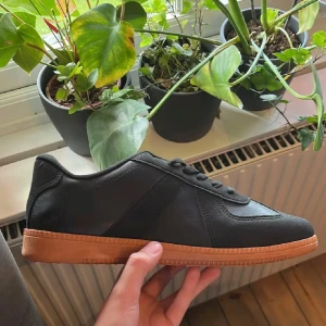Maison margiela  - Stilrena svarta sneakers med klassisk design och brun gummisula. Skorna har snörning och rund tå, tillverkade i ett material som liknar skinn. Perfekta för dig som gillar en clean och tidlös look.