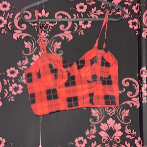 Röd och svart rutig korsettopp - Cool röd och svart rutig korsettopp med smala axelband och croppad passform. Toppen har en markerad byst och är perfekt för att sticka ut med sitt rutiga mönster. Passar dig som gillar edgy och trendiga plagg.