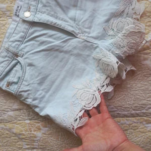 Ljusblå jeansshorts med spetsdetaljer - Supersnygga ljusblå jeansshorts från Denim med unika vita spetsdetaljer i blommigt mönster längs bensluten. Klassisk femficksmodell och knappstängning framtill. Perfekta för sommarens alla äventyr och ger en romantisk touch till din outfit.