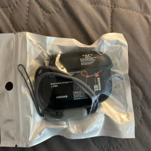 AirPods skal  - Säljer en svart AirPods skal med tryck och textdetaljer i vitt och orange. Skalet har ett smidigt snöre för att bära runt handleden eller hänga på kläderna exempelvis byxor. Perfekt för hörlurar.