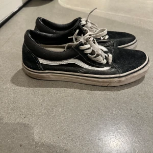 Svarta Vans Old Skool sneakers - Klassiska Vans Old Skool sneakers i svart med vit sula och vit sidostrip. Skorna har snörning, rund tå och är tillverkade i mocka och syntet. Perfekta för en avslappnad streetstil och funkar året runt.
