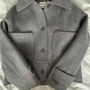 Grå ullrock från Kappahl XS - Snygg grå rock från Kappahl i storlek XS. Jackan har klassisk krage, stora framfickor och stängs med knappar. Säljer billigt då den inte är min stil längre, inga defekter alls!!