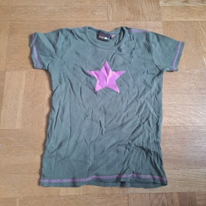 Grön t-shirt med rosa stjärna  - Cool grön t-shirt från Me&i med en stor rosa stjärna på bröstet och matchande rosa sömmar. Klassisk rund halsringning och korta ärmar. Perfekt för dig som gillar färg och vill sticka ut lite extra. Barnstorlek men kan passa xs, eventuellt s