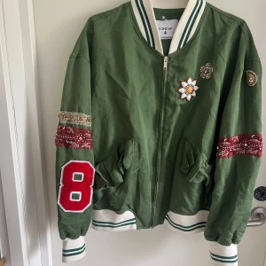 Grön bomberjacka från Dondup - Grön bomberjacka från Dondup med vita och gröna ribbade muddar, broderade blommor och pärldetaljer på bröstet, samt röda bandana-detaljer och en stor röd 8:a på ärmen. Jackan har dragkedja och två stora fickor framtill. Storlek 44 men är som en svensk storlek 40