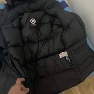 Svart dunjacka från Canada Goose - Säljer en svart dunjacka från Canada Goose med klassisk patch på ärmen och fet pälskant på huvan. Jackan är rejält vadderad och har dragkedja framtill. Perfekt för kalla vinterdagar och riktigt snygg streetstil . Jackan är äkta 