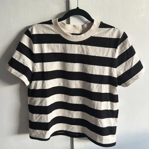 Randig t-shirt från Lindex, svart/vitbeige - Snygg svartvit randig t-shirt från Lindex i storlek M. Kortare och tight modell  med korta ärmar och rund hals. Tillverkad i återvunnen bomullsblandning, perfekt för en chill och stilren look. Snygga till jeans. Ny, endast provad. Ingen prislapp kvar. 