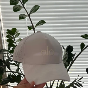 Vit keps från Alo med meshdetaljer - Snygg vit keps från Alo med böjd skärm och ventilerande meshpaneler på sidorna. Kepsen har justerbar rem bak och diskret Alo-logga framtill. Perfekt för träning eller soliga dagar. Tillverkad i lätt syntetmaterial för maximal komfort.