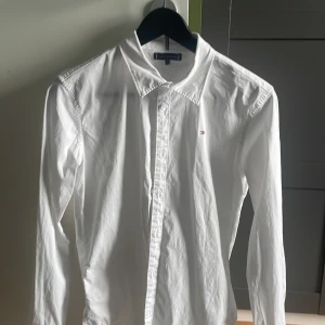 Vit skjorta från Tommy Hilfiger  - Klassisk vit skjorta från Tommy Hilfiger i storlek 164. Skjortan har lång ärm, knappar framtill och en liten broderad logga på bröstet. Tillverkad i mjuk bomull och har en stilren, tidlös design som passar till många olika looks.