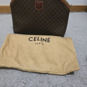 Brun väska från Celine Paris - Säljer en brun väska från Celine Paris med klassiskt monogrammönster och läderdetaljer. Väskan har en rundad form upptill, dragkedja och insida i ljusbrunt skinn. Perfekt för dig som gillar vintage och lyxiga accessoarer.