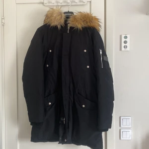 Svart parkas Rockandblue XL med fuskpäls - Säljer en svart parkas från Rockandblue i storlek XL. Jackan har en stor huva med snygg fuskpäls, flera fickor med silverfärgade knappar och dragkedja. Perfekt för kalla dagar med sin långa modell och varma polyesterfoder.