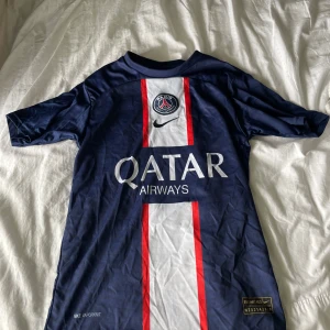 PSG Mbappé matchtröja Nike 164 - Paris Saint-Germain fotbollströja med Mbappé 7 på ryggen. Mörkblå med vit och röd rand längs mitten, broderat PSG-märke och Nike-logga. Qatar Airways sponsortryck. Tillverkad i Dri-FIT material som andas, perfekt för fotbollsträning eller att bära på läktaren.
