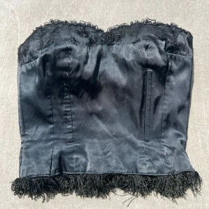 Svart korsett med spets och fransar - Snygg svart korsett med spetsdetaljer upptill och fransar längs nederkanten. Axelbanden är röda men jag brukar stoppa in dem så att korsetten är axelbandslös. Stängs med hyskor framtill och har en glansig satinliknande yta. 