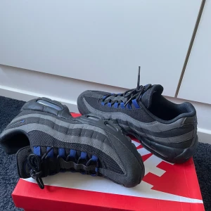 Nike Air Max 95 svart/blå sneakers - Nike Air Max 95 i svart och djupblå med grå detaljer. Ovandel i mesh och mocka, klassisk vågig design och synlig Air-dämpning i sulan. Svarta snören och blå öglor ger en sportig vibe. Perfekta för dig som gillar streetstyle och komfort.