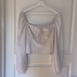 Vit blus med knyt och puffärm - Supersöt vit blus med långa puffärmar och croppad passform. Toppen har en fyrkantig ringning och knyt fram, samt ett strukturerat tyg som ger en extra detalj. Perfekt för dig som gillar sommrig och enkel men detaljrik still. 