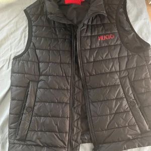 Svart dunväst från Hugo Boss - Snygg svart quiltad dunväst från Hugo Boss med röd HUGO-logga på bröstet. Västen har dragkedja framtill, två fickor med knappstängning och en hög krage. Perfekt för lager-på-lager och ger en clean streetstyle-vibe.