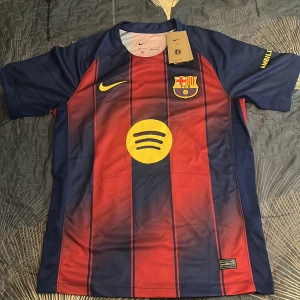 FC Barcelona Nike matchtröja blå/röd - FC Barcelona matchtröja från Nike med klassiska blå och röda vertikala ränder, klubbmärke på bröstet och gul Nike-logga. Tröjan har korta ärmar, rund hals och är tillverkad i lätt, ventilerande funktionsmaterial. Spotify och UNHCR-loggor i gult tryck på fram- och baksidan.