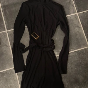 Svart långärmad klänning med bälte - Svart långärmad klänning med markerad midja och bälte med snyggt spänne. Klänningen har en smickrande passform och är tillverkad i ett ribbat material som ger en cool struktur. Perfekt för dig som gillar stilrena och trendiga plagg.