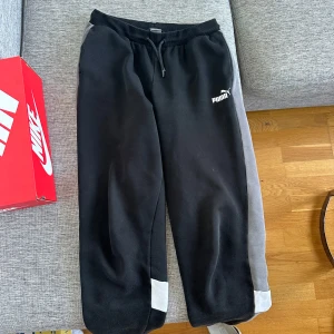 Svarta och grå mjukisbyxor från Puma - Snygga svarta mjukisbyxor från Puma med gråa paneler längs sidorna och vita detaljer vid bensluten. Byxorna har resår i midjan med snörning och Puma-logga på låret. Perfekta för chill eller träning.