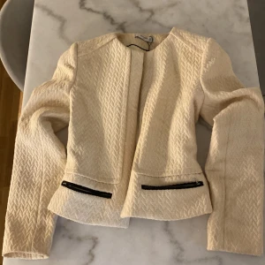 Beige kavajjacka från Mango XS - Snygg beige kavajjacka från Mango i storlek XS. Jackan har ett strukturerat mönster, raka axlar och två svarta dragkedjor framtill. Materialet känns mjukt och har en lätt quiltad känsla. Perfekt för dig som gillar stilrena och trendiga plagg.