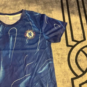 Chelsea blå fotbollströja herr - Chelsea fotbollströja i blått med klubbens logga på bröstet. Tröjan har korta ärmar och ett unikt abstrakt mönster i olika blå nyanser. Tillverkad i lätt och ventilerande polyester, perfekt för match eller träning.