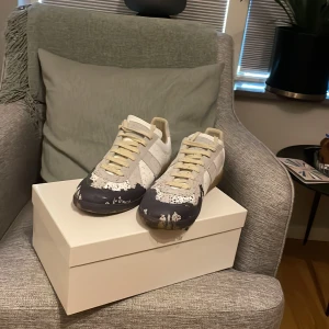 Maison Margiela sneakers med färgstänk - Unika sneakers från Maison Margiela i vitt och grått skinn med beige snörning och coolt svart färgstänksmönster på tån och sidorna. Klassisk siluett med gummisula och diskreta detaljer som ger en edgy vibe. Perfekta för dig som vill sticka ut.