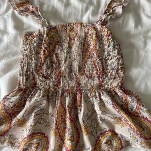 Beige paisleymönstrad linnetopp - Supersöt linnetopp i beige med paisleymönster i rött, gult och vitt. Smockad överdel och volangig nederdel, samt breda axelband. Perfekt för varma dagar och ger en bohemisk vibe. Märke är okänt