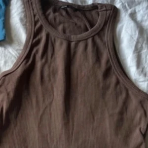 Brun och svart ribbade linnetoppar - Två stilrena linnetoppar, en i brun och en i svart. Båda är ribbade och har hög halsringning samt ärmlöst snitt. Perfekta att matcha med jeans eller kjol för en clean look. Säljes tillsammans eller separat.