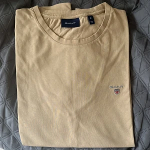 GANT T-SHIRT - BEIGE - STORLEK M - Snygg beige t-shirt från GANT med klassisk rund hals och broderad logga på bröstet. Tillverkad i mjuk bomull som känns skön mot huden. Enkel och stilren design som funkar till det mesta. Perfekt för dig som gillar clean och tidlös stil. Äkta vara köpt ifrån Boozt. (Tröjan innehåller ett litet stickhål längst ner på tröjan vid magen, inget synligt men dock sänkt pris på grund av det, 200-150 istället)