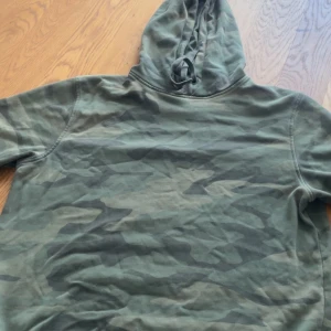 Grön camo hoodie från Etirel - Säljer en grön hoodie från Etirel i camouflagemönster. Tröjan har huva med snörning och är tillverkad i mjukt bomullsmaterial. Perfekt för dig som gillar streetwear och vill ha en chill look.