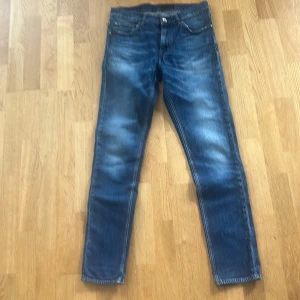 Blå jeans från Tiger of Sweden - Snygga blå jeans från Tiger of Sweden med klassisk femficksdesign och orangea sömmar. Jeansen har en smal passform och raka ben, tillverkade i mjukt jeanstyg. Perfekta för dig som gillar stilrena och tidlösa jeans. Storleken är 32/34.