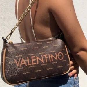 Brun axelväska från Valentino - Snygg brun axelremsväska från Valentino med logotypmönster och guldkedja * köpt från zalando för 1 095. Väskan har en avtagbar axelrem i beige och dragkedja (2 olika band medföljer). Mycket bra skick med inga defekter. Höjd: 14 cm i storlek One Size Längd: 25 cm i storlek One Size Vidd: 5 cm i storlek One Size