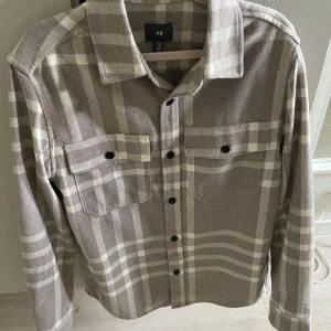 Grå Beige rutig skjortjacka från hm - Snygg beige och grå skjortjacka från H&M i rutig design. Skjortan har två bröstfickor med lock och svarta knappar framtill. Mjuk och skön känsla, passar till många outfits. Knappt använd 