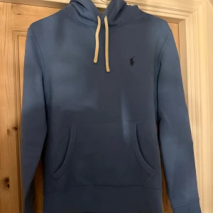 Blå hoodie från Ralph Lauren - Snygg blå hoodie från Ralph Lauren med klassisk broderad logga på bröstet. Tröjan har huva med vita snören och en stor magficka framtill. Perfekt för dig som gillar en enkel och stilren tröja. Strl Xs passar S/M. Långärmad och i mjuk bomull. Inga defekter över huvud taget, knappt använd då den var för liten. Tveka inte att höra av er! Kan sänka priset vid snabb affär!              