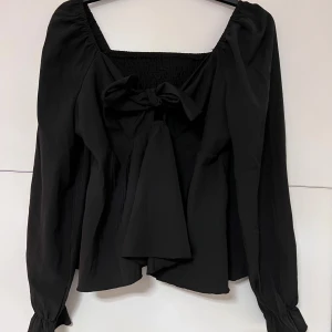 Svart blus med rosett och smock - Supersöt svart blus med lång ärm, smockad överdel och stor rosett framtill. Blusen har en fyrkantig ringning och volangiga ärmslut. 